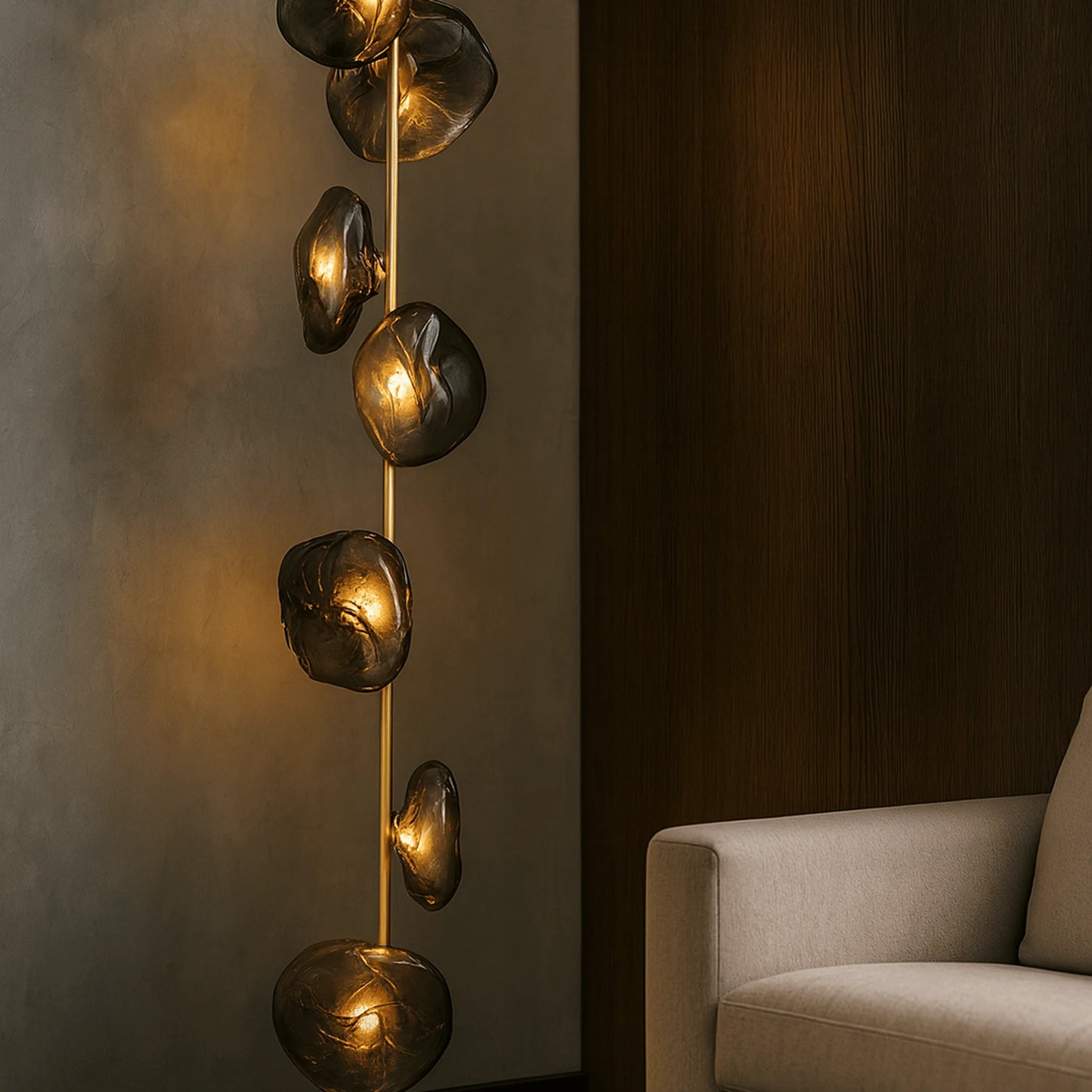 Modern LED floor lamp with a gold stem and organic, smoky grey glass shades emitting a warm glow, positioned next to a contemporary armchair.