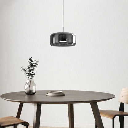 A modern smoked glass pendant light hangs above a round wooden dining table featuring a glass vase with greenery and stacked plates in a minimalist room.