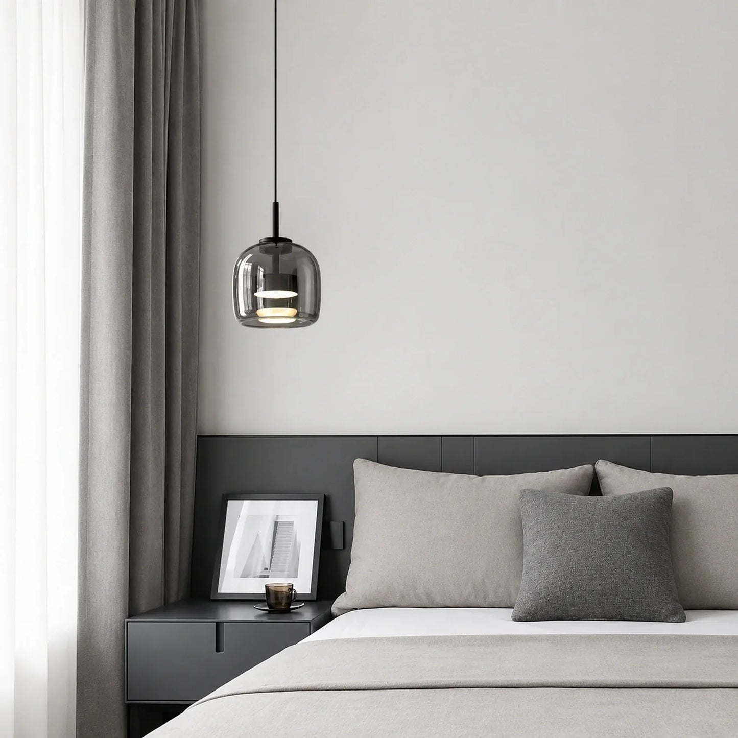 Modern glass pendant light with a smoked finish hanging above a minimalist bedside table in a contemporary bedroom with grey bedding and curtains.