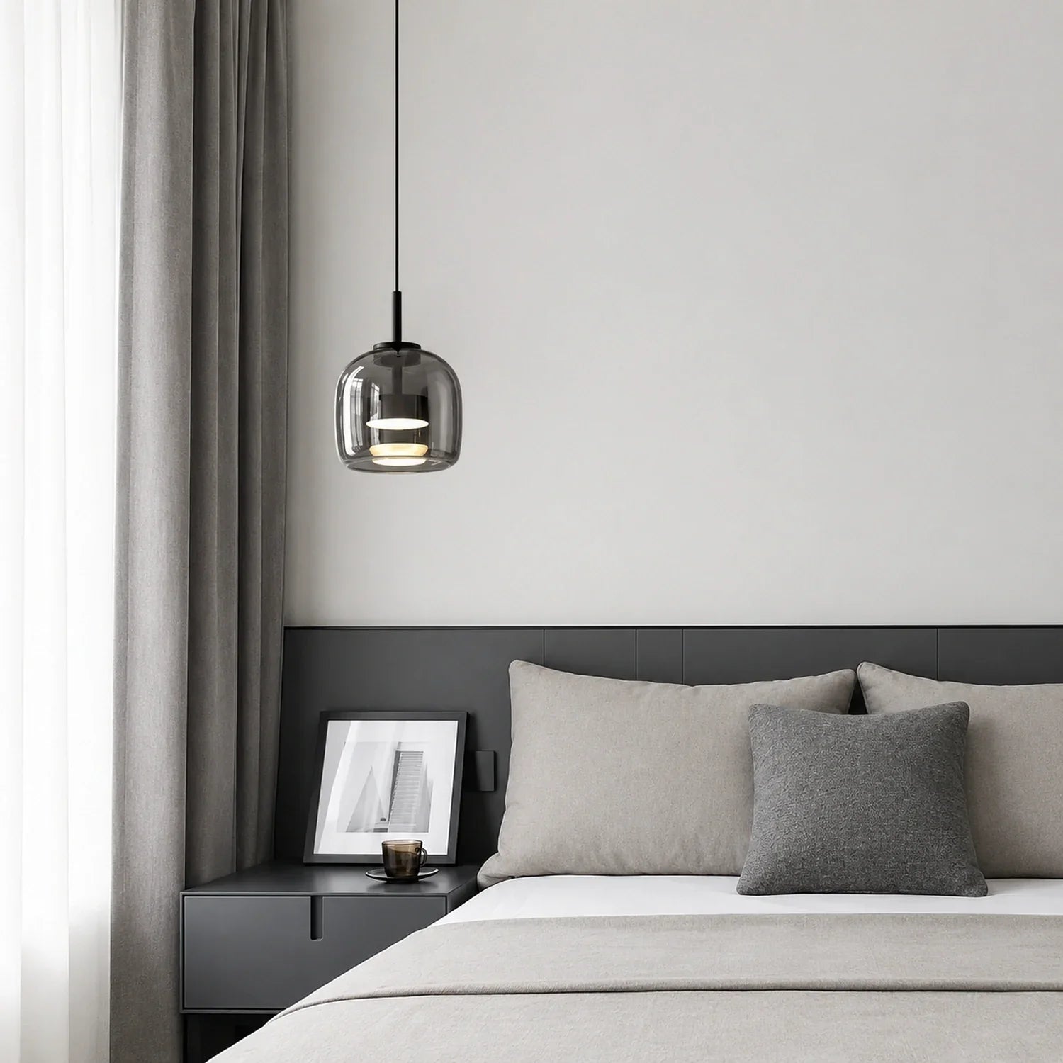 Modern glass pendant light with a smoked finish hanging above a minimalist bedside table in a contemporary bedroom with grey bedding and curtains.