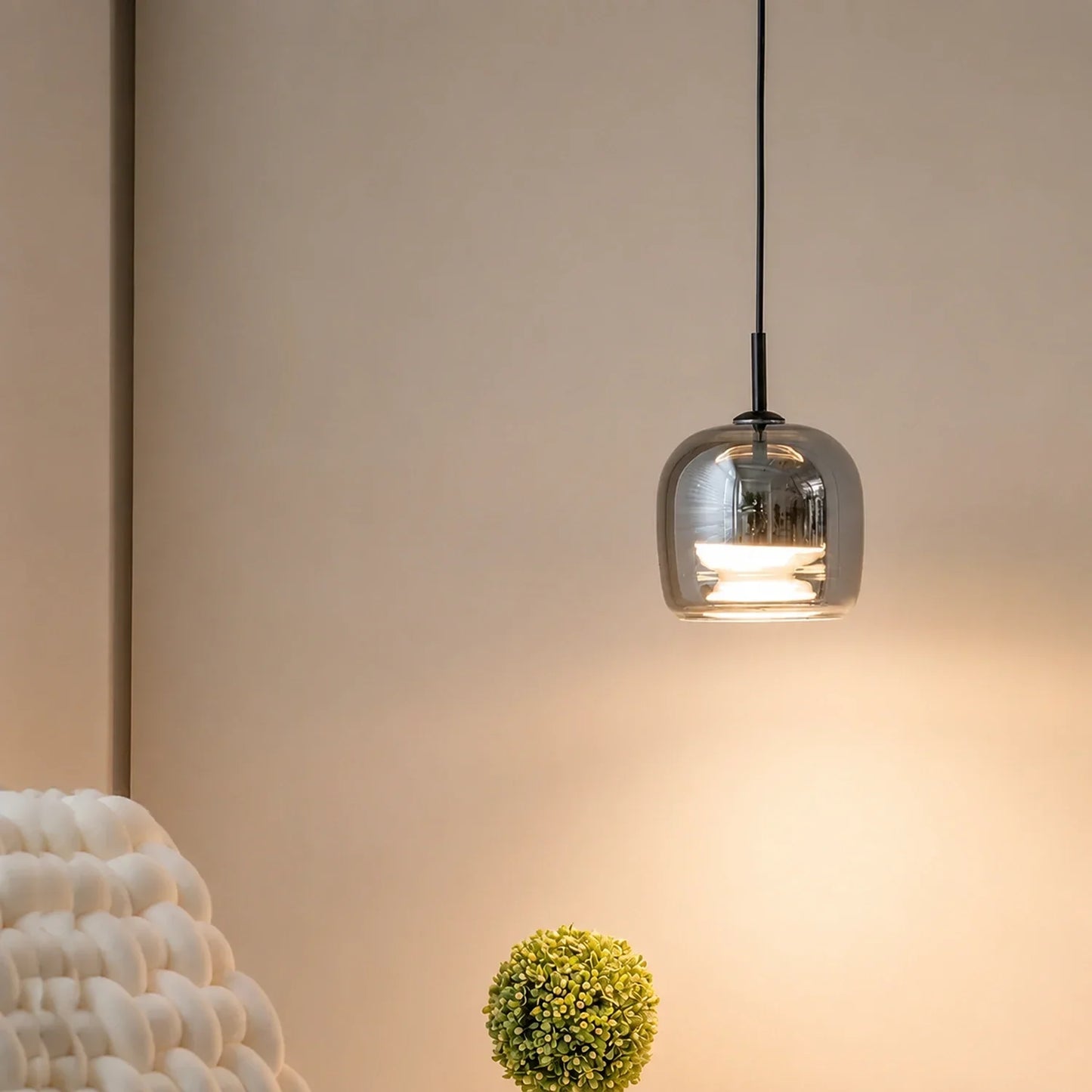A modern pendant light with a sleek, smoked glass shade hanging from a black cable, casting a warm glow against a neutral wall in a minimalist room.
