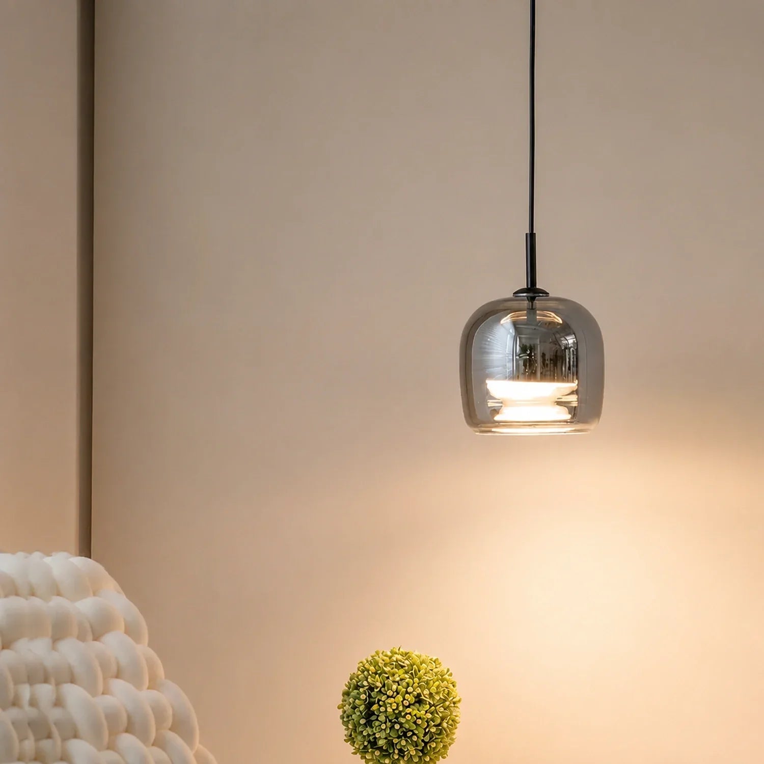 A modern pendant light with a sleek, smoked glass shade hanging from a black cable, casting a warm glow against a neutral wall in a minimalist room.