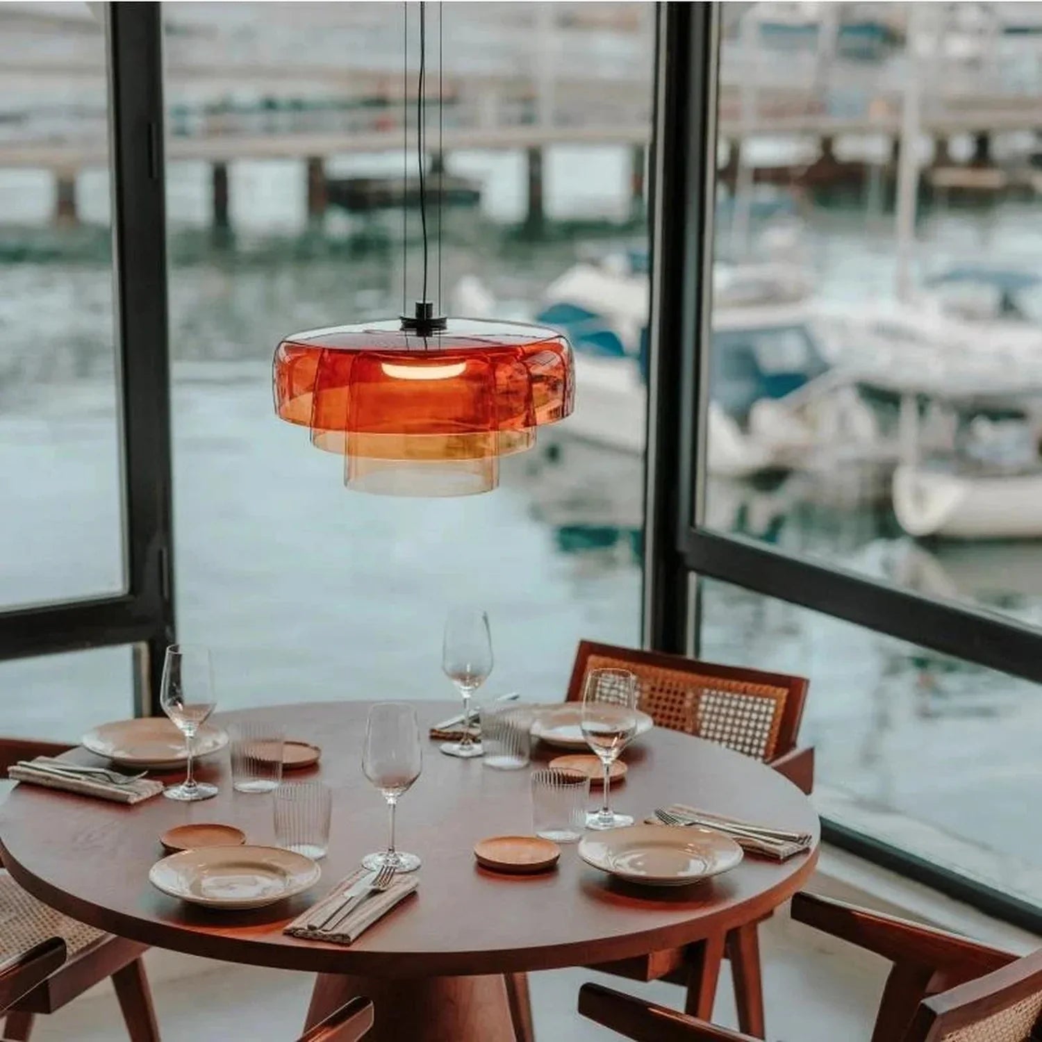 A modern amber glass pendant light with integrated LED hanging above a circular wooden dining table in a room overlooking a marina.