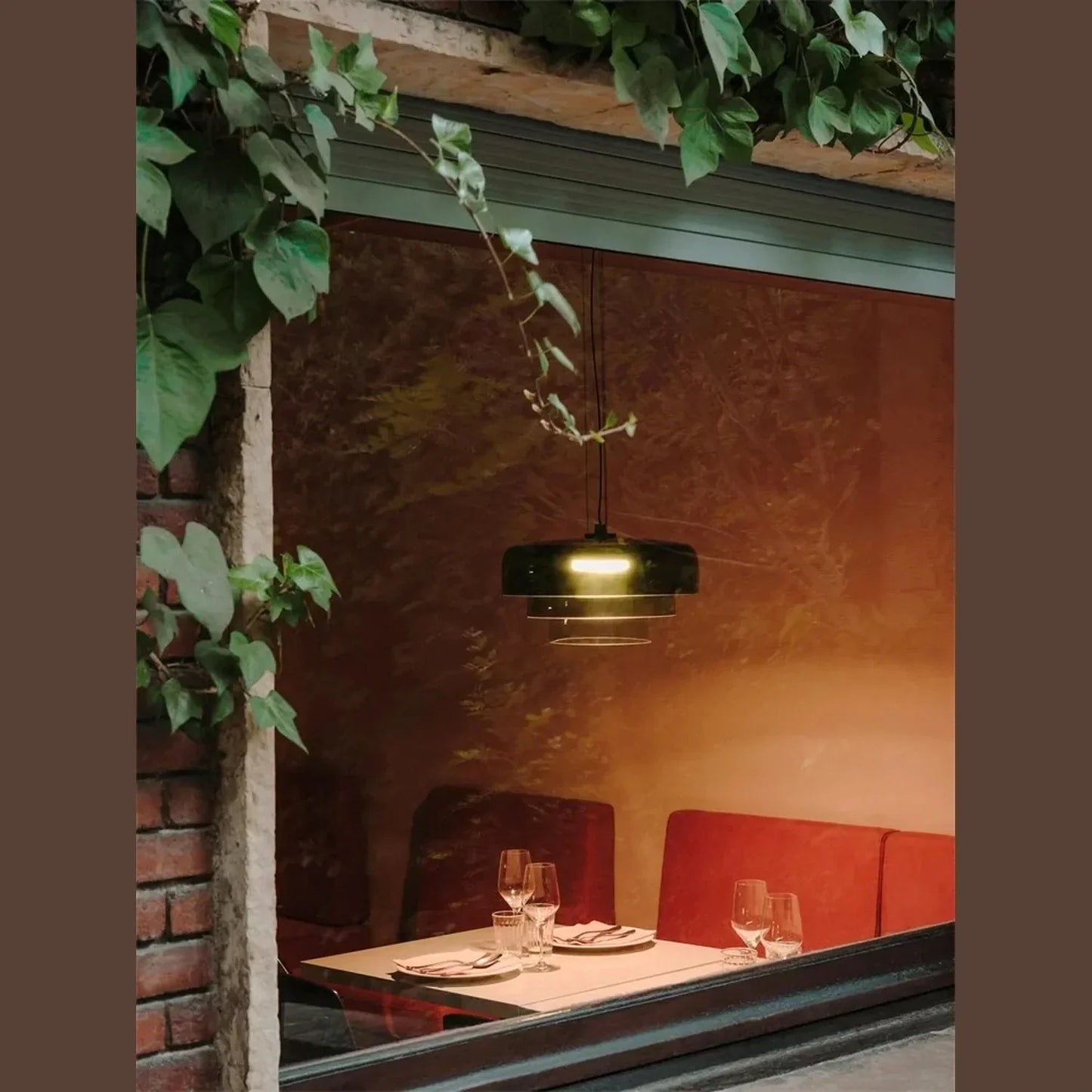 Modern tiered glass pendant light with integrated LED illuminating a cosy restaurant dining table seen through a window framed by climbing ivy.