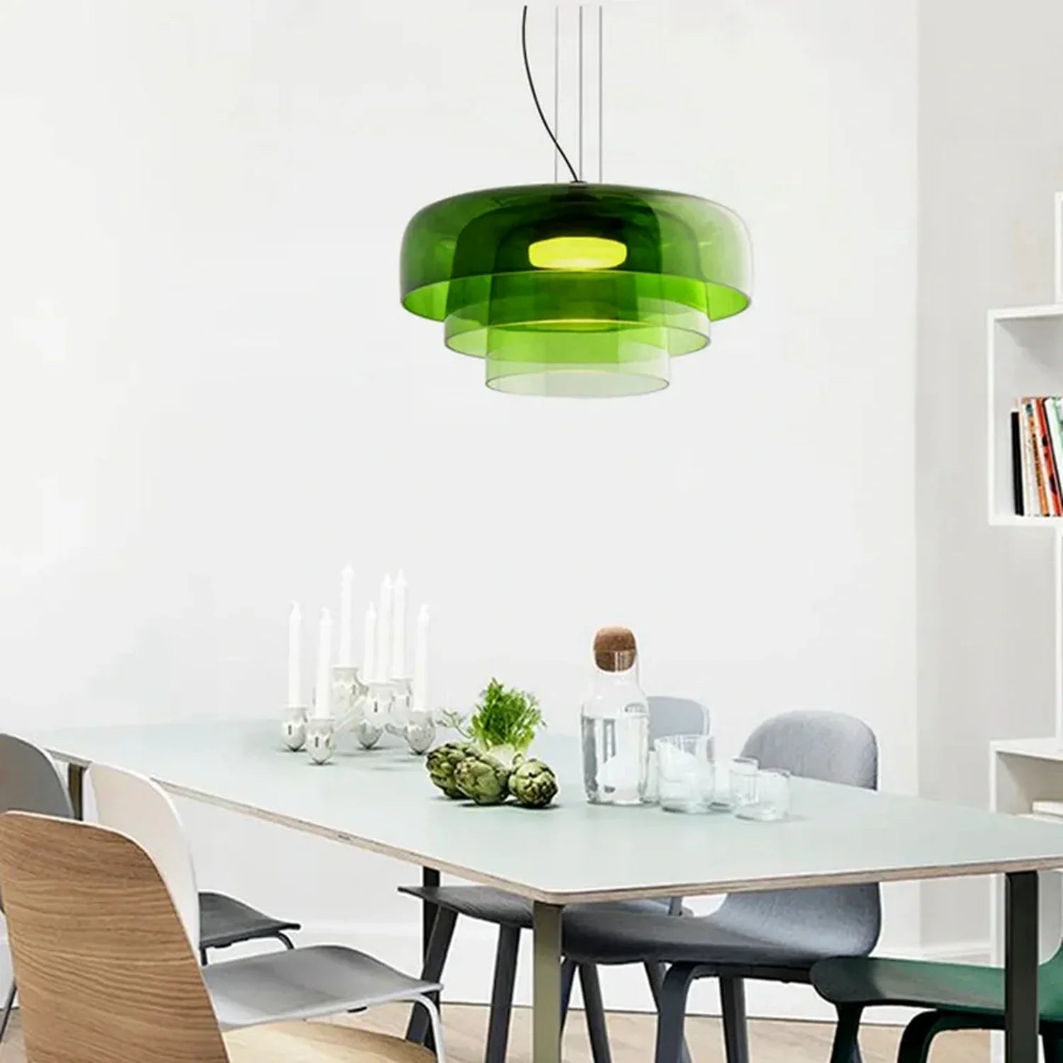 A modern tiered green glass pendant light with integrated LED, suspended above a contemporary dining table set with candles and fresh artichokes.
