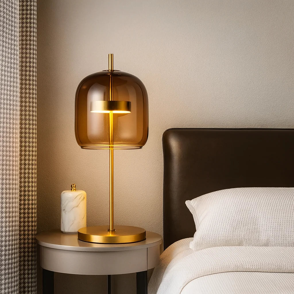 Modern table lamp with a smoked glass dome shade and brushed gold metal base, glowing warmly on a bedside table next to a bed and marble accessory.