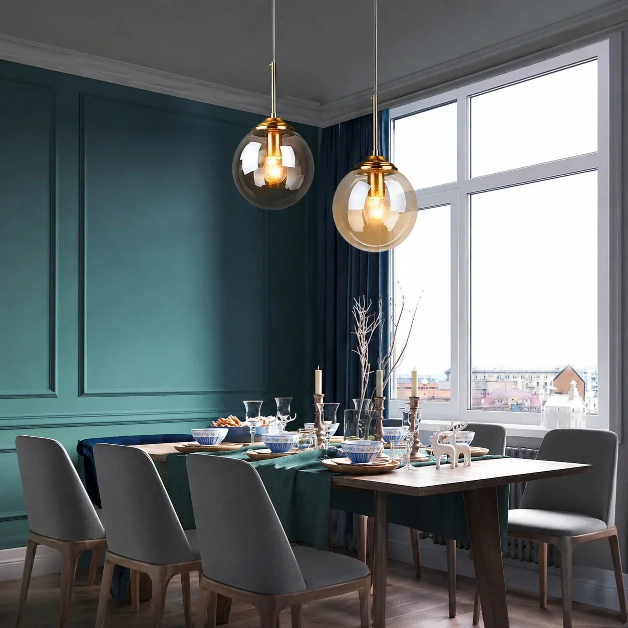 Two modern globe glass pendant lights with gold fittings hanging above a wooden dining table in a stylish room with teal panelled walls and large windows.