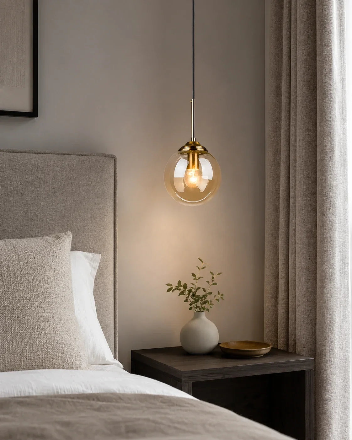 Modern globe glass pendant light with brass detailing hanging above a bedside table in a neutral bedroom, providing warm ambient lighting.