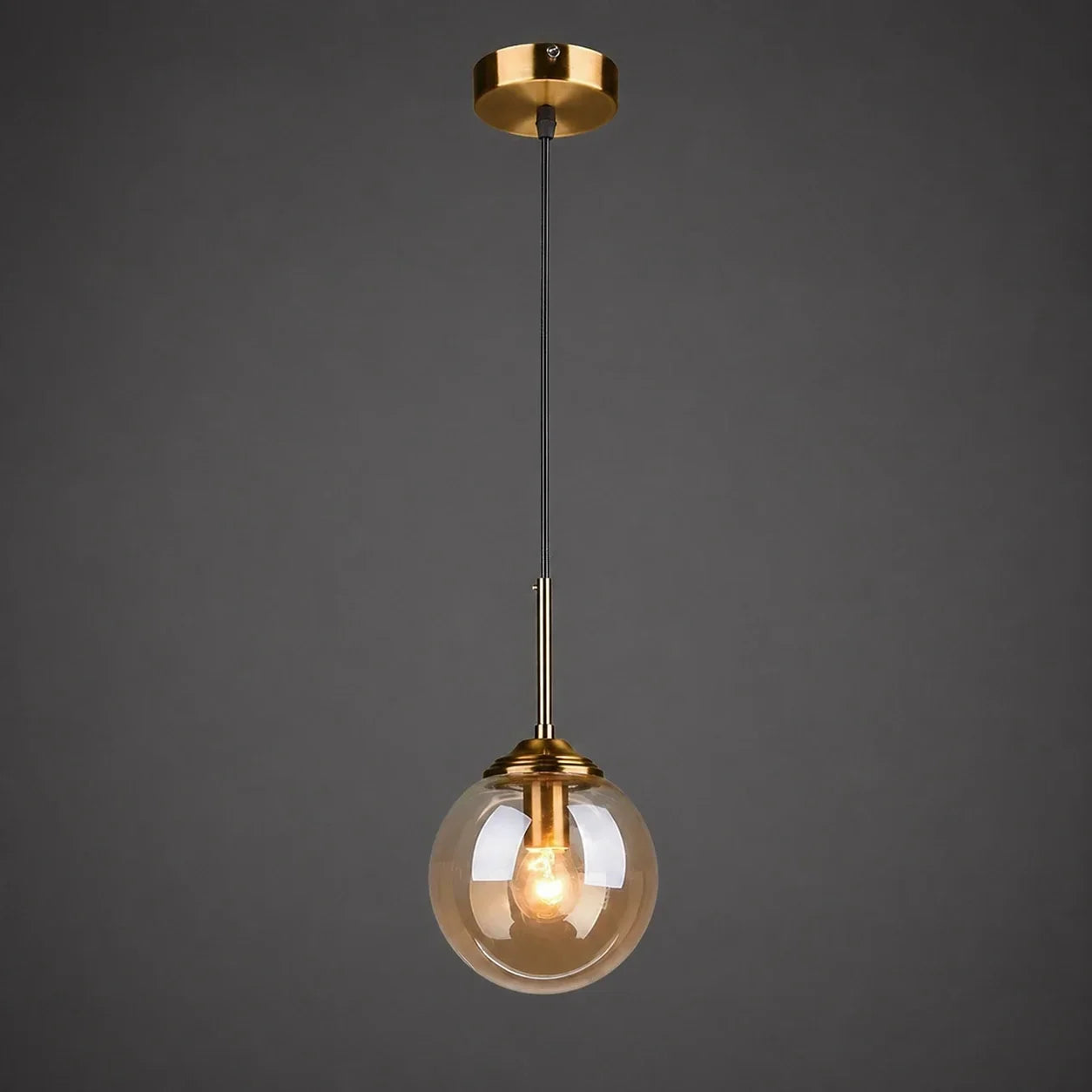 Modern globe pendant light featuring an amber glass shade and brushed brass finish, suspended by a black cable against a dark grey background.