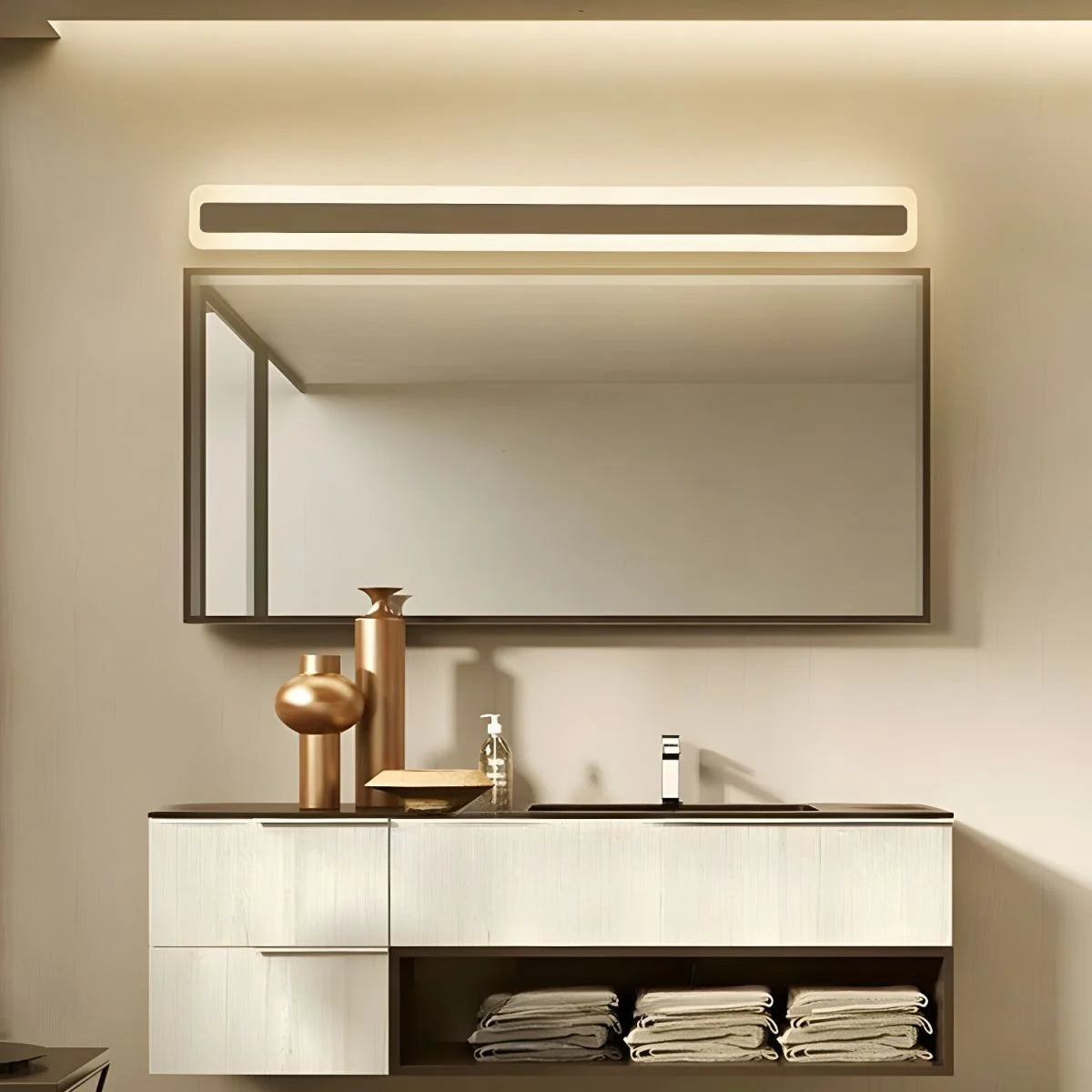 A modern gold LED wall light mounted above a rectangular bathroom mirror, illuminating a vanity unit with stacked towels and decorative gold vases.