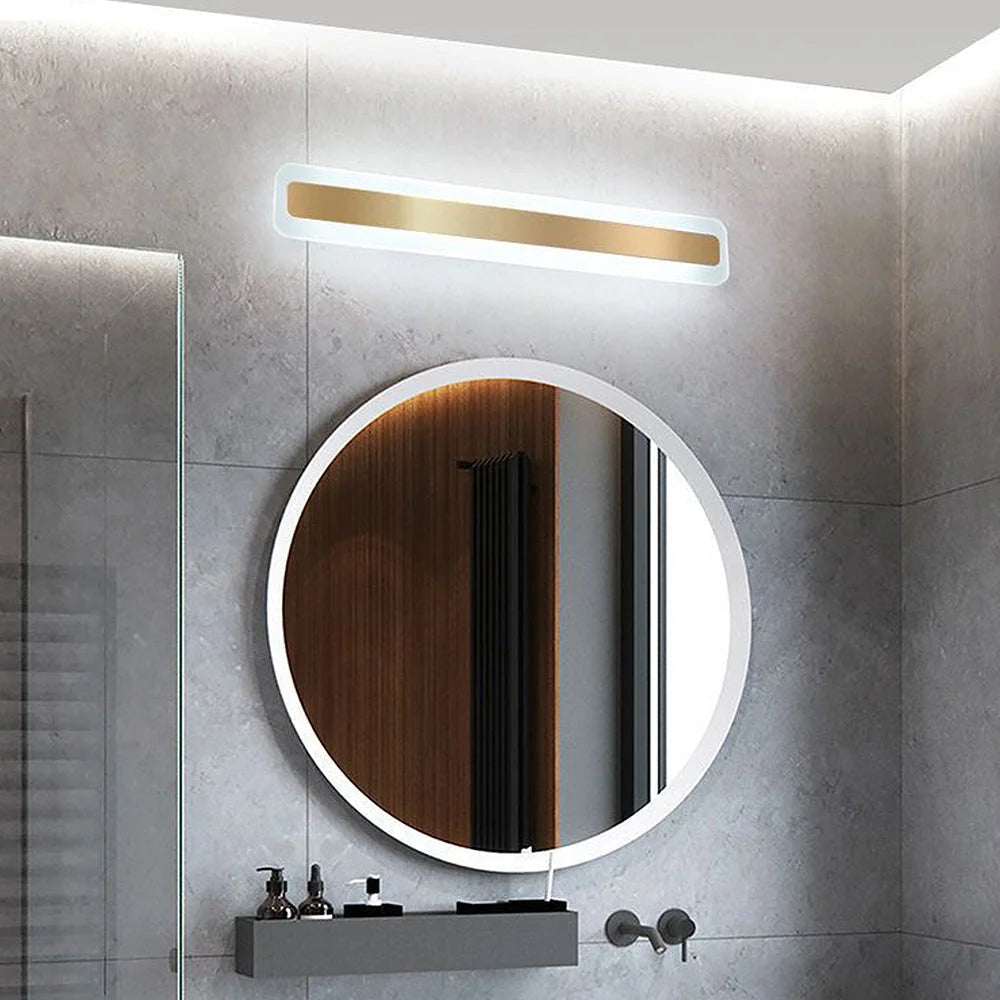 A modern, linear LED bathroom wall light with a gold finish mounted above a circular mirror in a contemporary grey tiled bathroom setting.