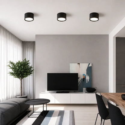 Three black surface-mounted aluminium LED downlights installed on a white ceiling in a modern, minimalist living room setting.