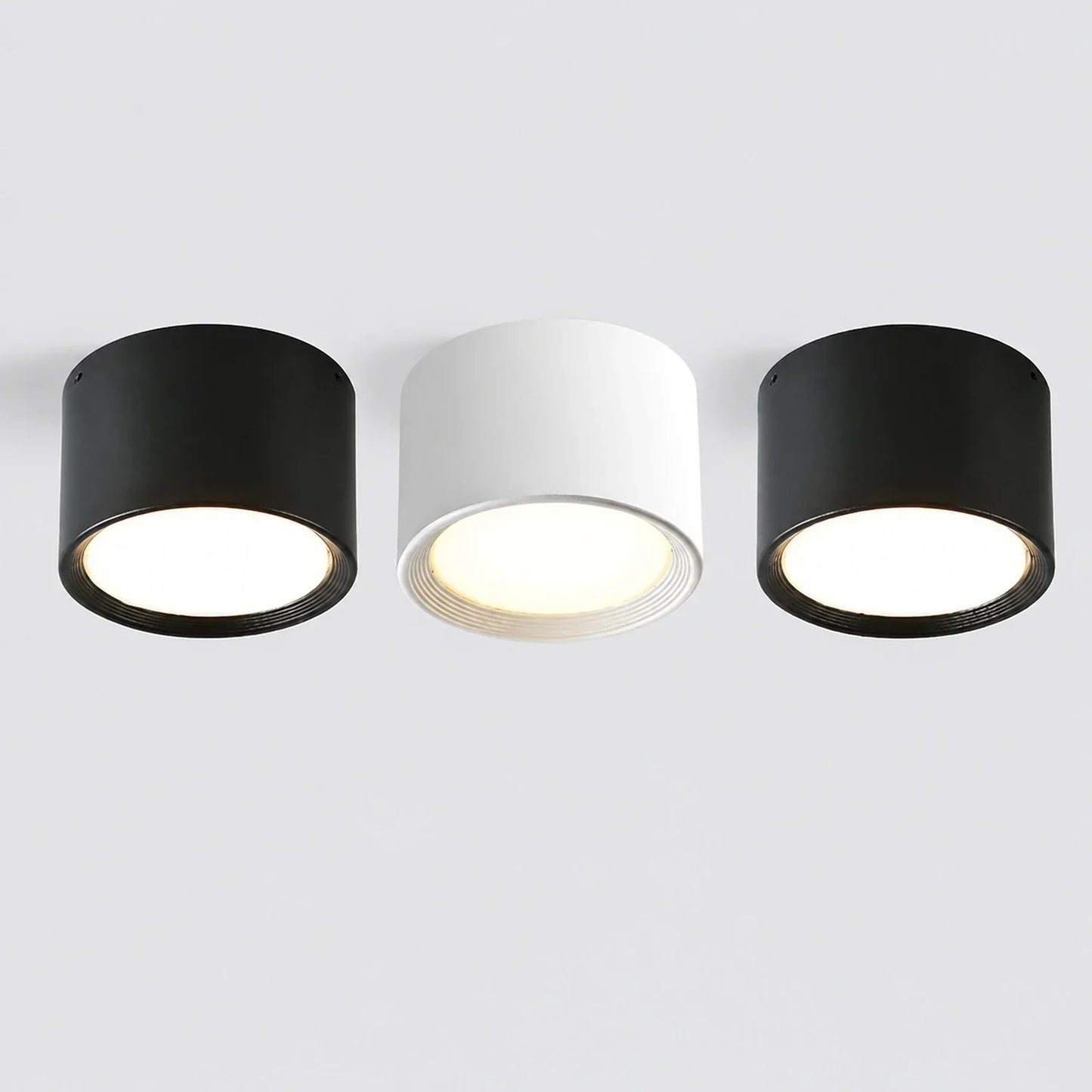 Three minimalist surface-mounted LED ceiling downlights in black and white, featuring a sleek cylindrical aluminium design with warm glowing light.