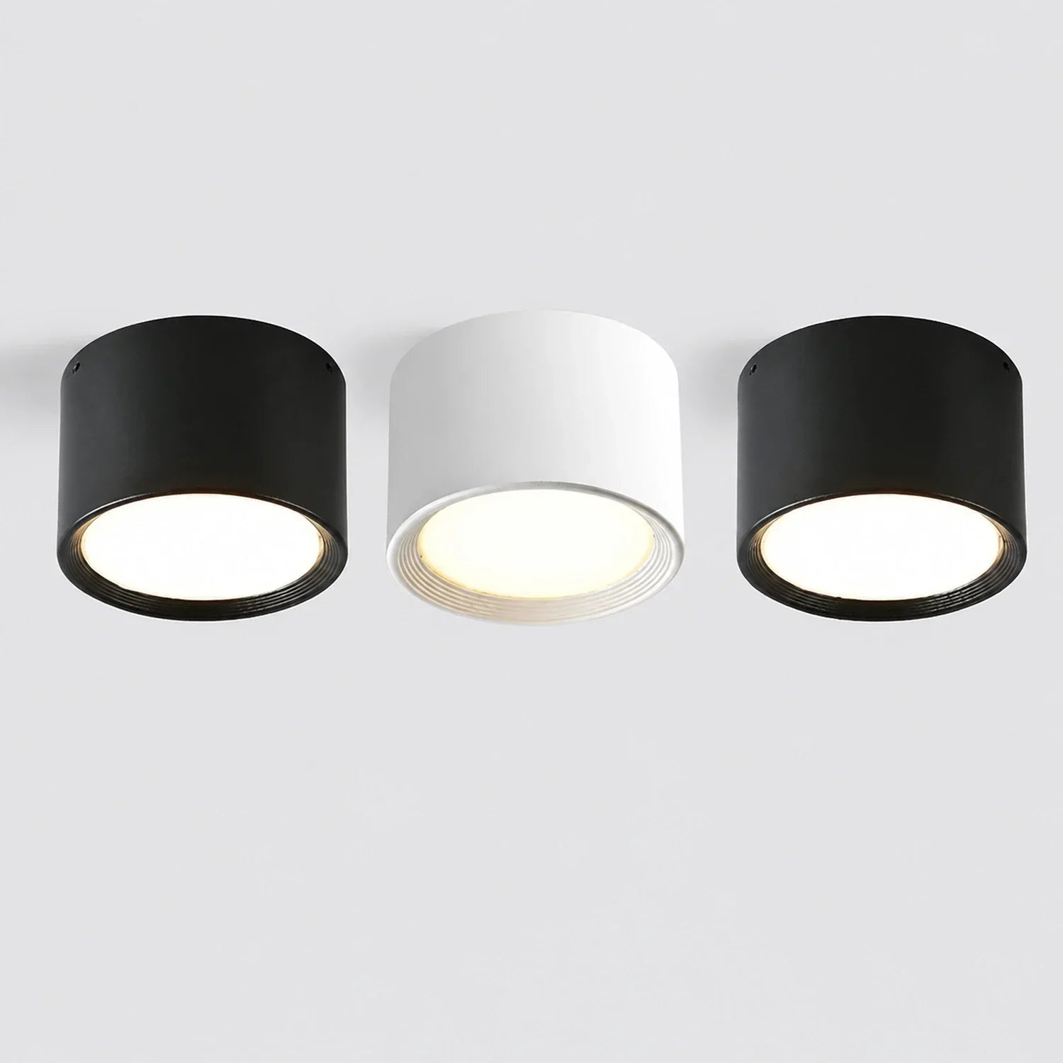 Three minimalist surface-mounted LED ceiling downlights in black and white, featuring a sleek cylindrical aluminium design with warm glowing light.