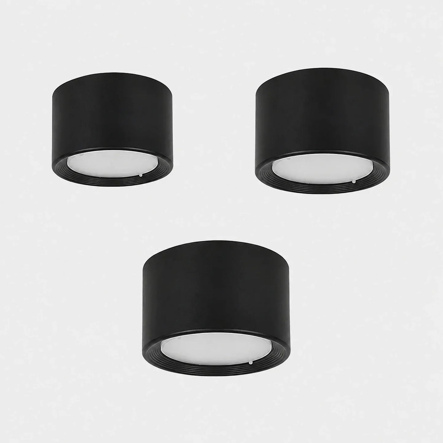 Three minimalist black cylindrical surface-mounted LED ceiling downlights with white diffusers, arranged on a plain light grey background.