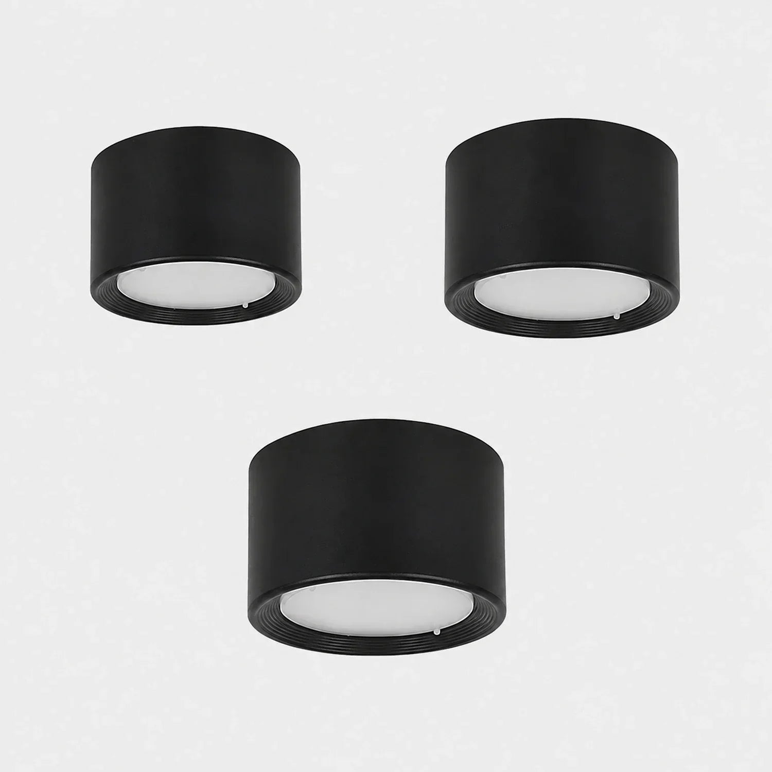 Three minimalist black cylindrical surface-mounted LED ceiling downlights with white diffusers, arranged on a plain light grey background.