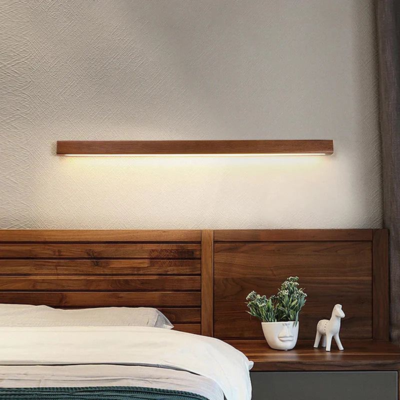 A modern linear wooden LED wall light casting a warm downward glow on a textured wall above a wooden bed headboard in a cosy bedroom.