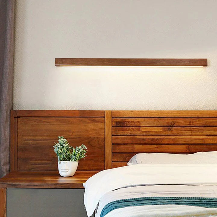 A modern linear wooden LED wall light emitting a warm downlight glow, mounted on a wall above a wooden headboard in a cosy bedroom.
