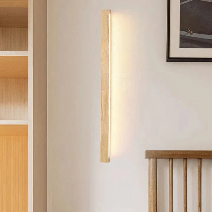 A modern vertical wooden LED wall light emitting a warm glow on a light-coloured wall next to a wooden headboard in a minimalist bedroom.