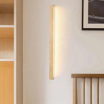 A modern vertical wooden LED wall light emitting a warm glow on a light-coloured wall next to a wooden headboard in a minimalist bedroom.