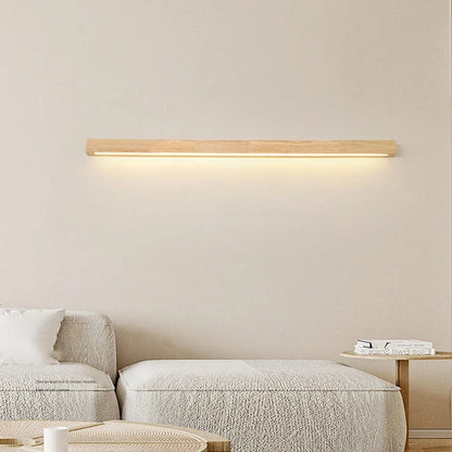 A modern linear wooden LED wall light emitting a warm downward glow, mounted on a neutral wall above a minimalist fabric sofa.
