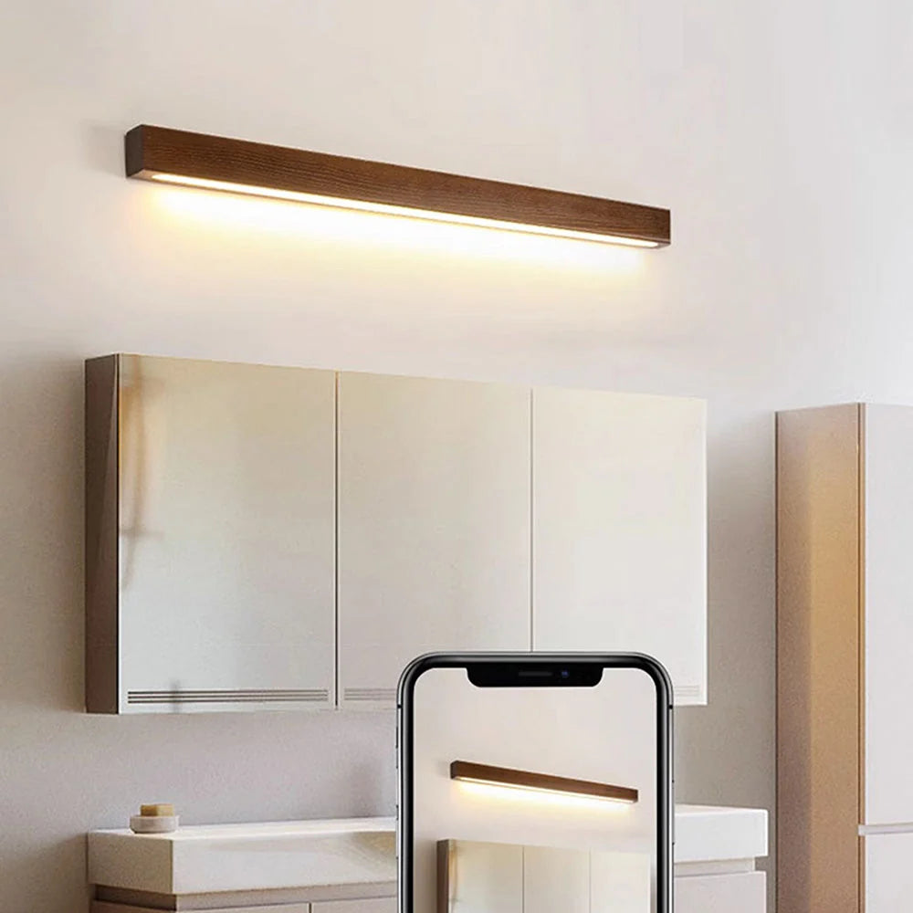 A modern linear wooden LED wall light emitting a warm glow above a mirrored bathroom cabinet, shown with a smartphone preview in the foreground.