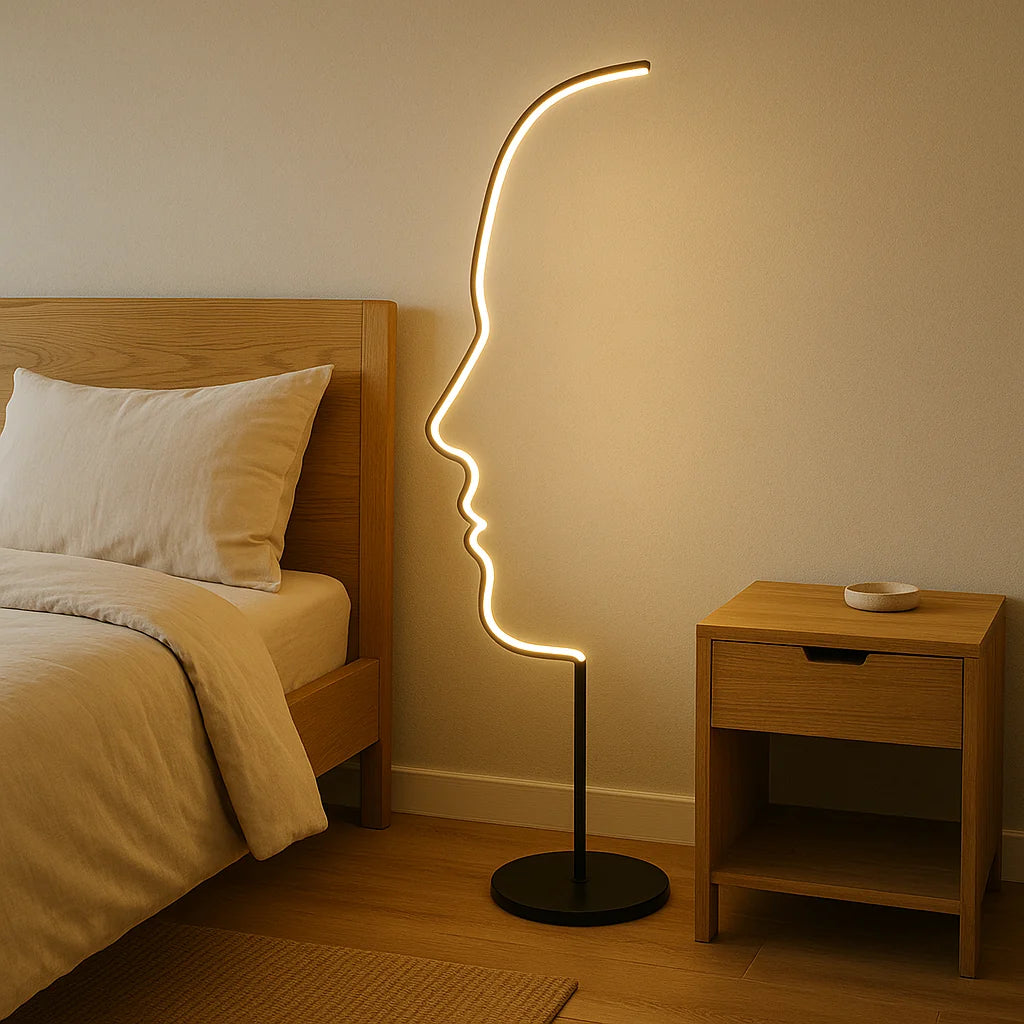 A modern LED floor lamp with an artistic human face profile design, glowing with warm light next to a wooden bed and bedside table in a bedroom.