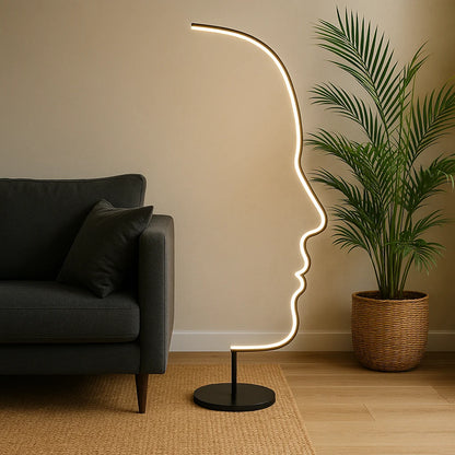 Modern LED floor lamp with an artistic human face outline design, glowing warmly next to a grey sofa and a potted palm in a living room.