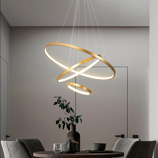 Modern gold three-ring LED ceiling light suspended above a dining table in a contemporary interior, featuring adjustable circular light fixtures.
