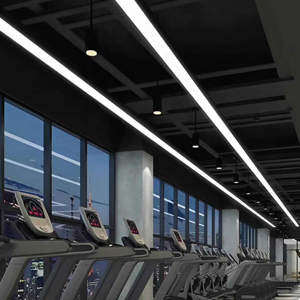 Modern linear LED pendant lights suspended from a dark ceiling in a commercial gym setting with rows of treadmills overlooking a city skyline at night.