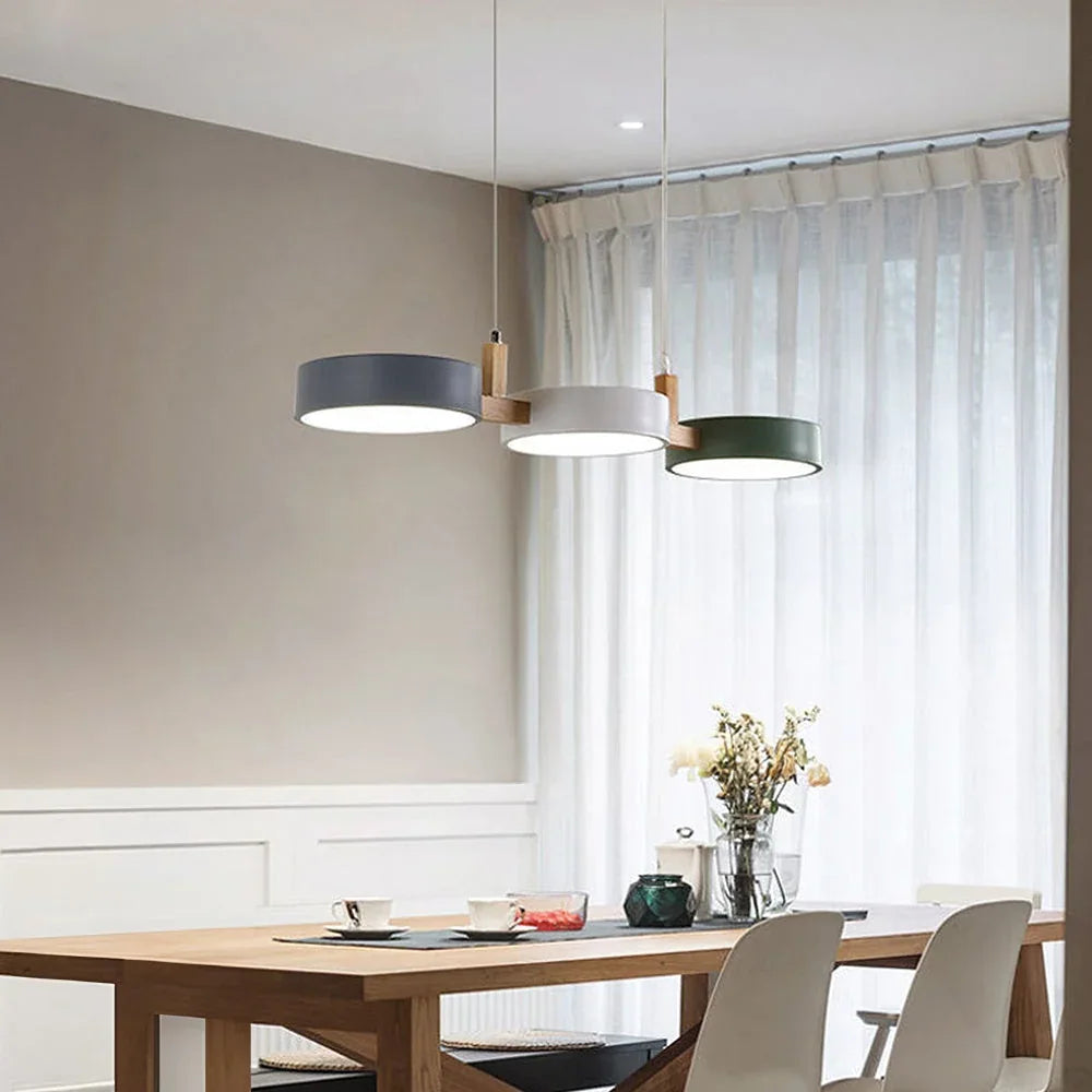 Modern sculptural 3-light pendant with grey, white and green circular shades and wood accents hanging above a wooden dining table in a bright room.