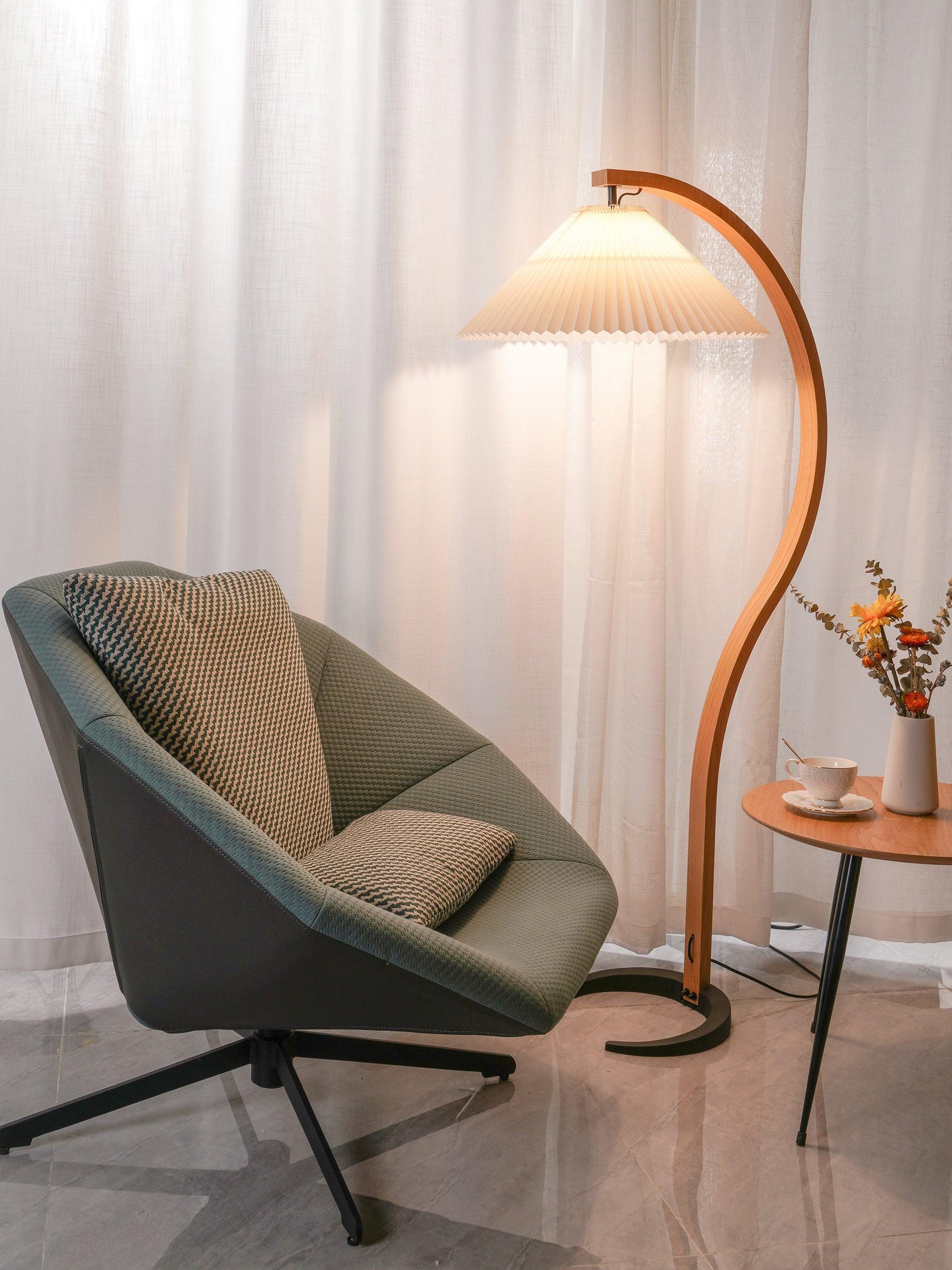 Nordic style beech wood floor lamp with a pleated shade illuminating a cosy corner featuring a modern green armchair and a small side table.