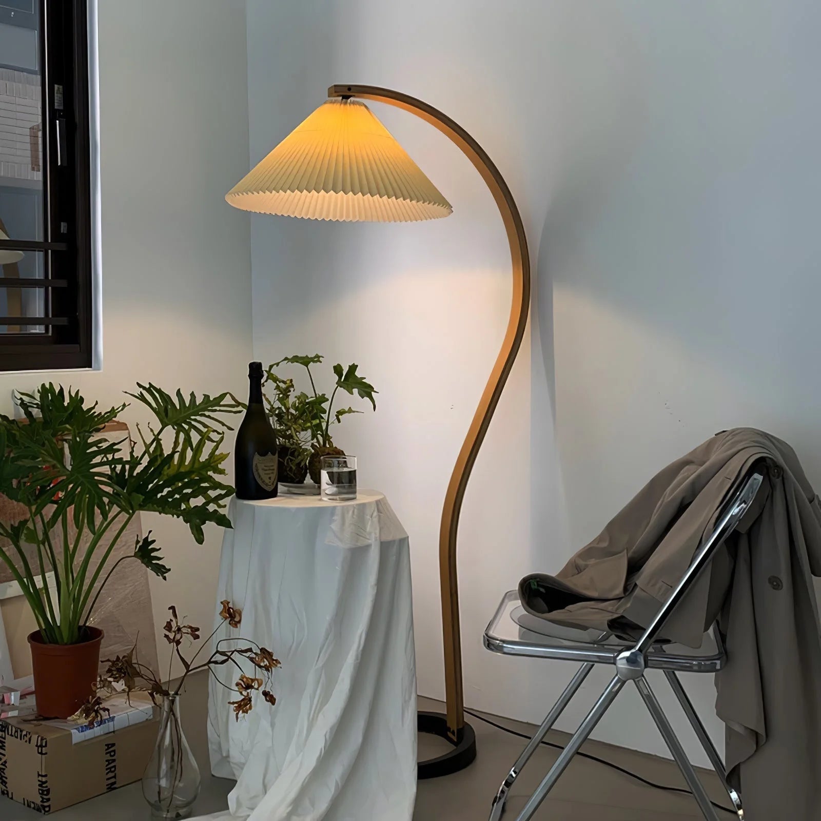 Nordic style floor lamp with a curved beech wood stand and a warm glowing pleated shade, positioned next to a small table and a modern metal chair.