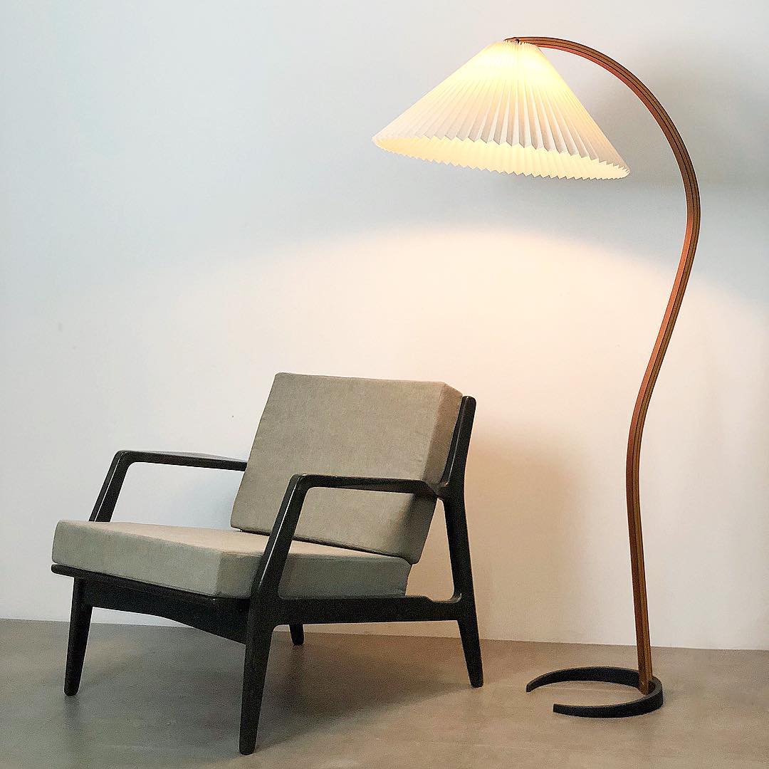 Nordic style floor lamp with a curved beech wood stand and a white pleated shade, illuminating a modern armchair in a minimalist living room setting.