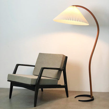 Nordic style floor lamp with a curved beech wood stand and a white pleated shade, illuminating a modern armchair in a minimalist living room setting.