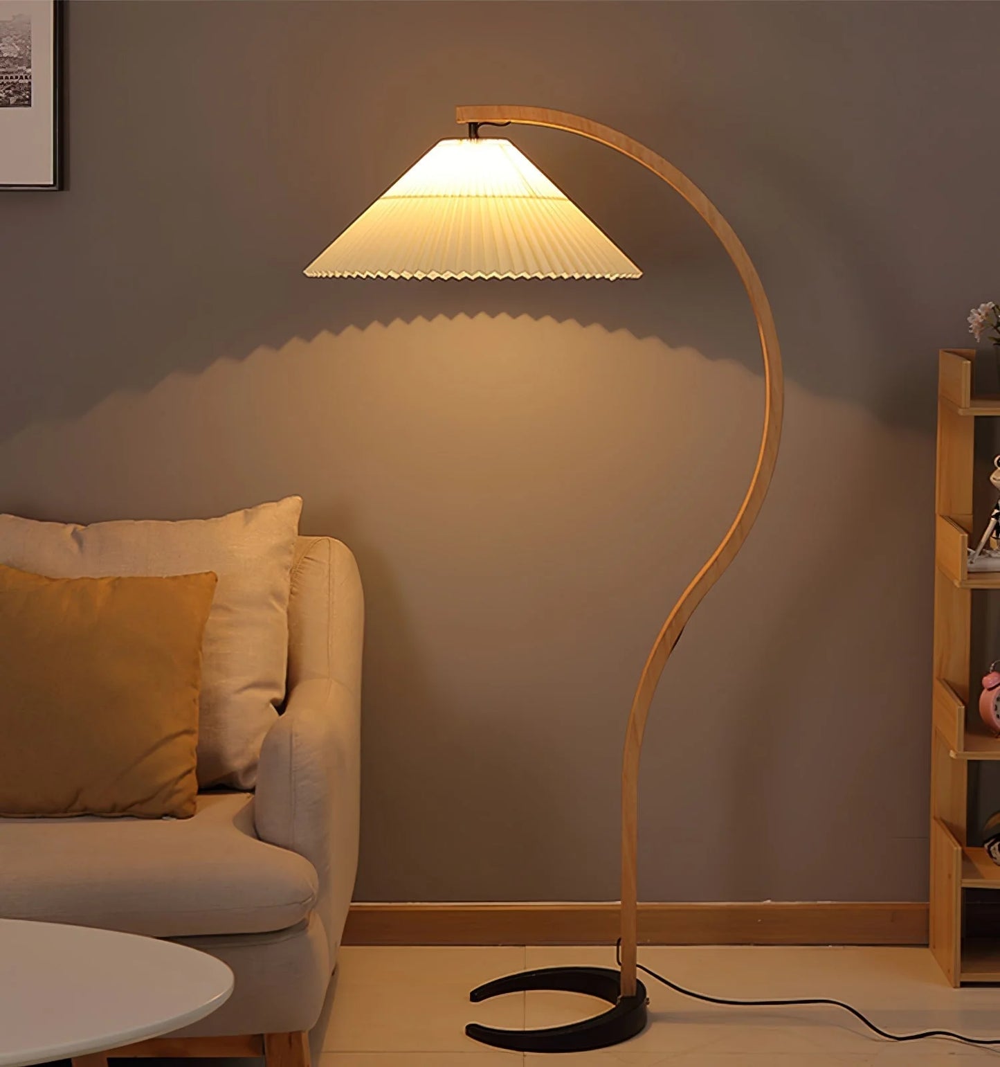 Nordic style floor lamp with a curved beech wood stand and a white pleated shade, casting a warm glow in a modern living room beside a neutral sofa.
