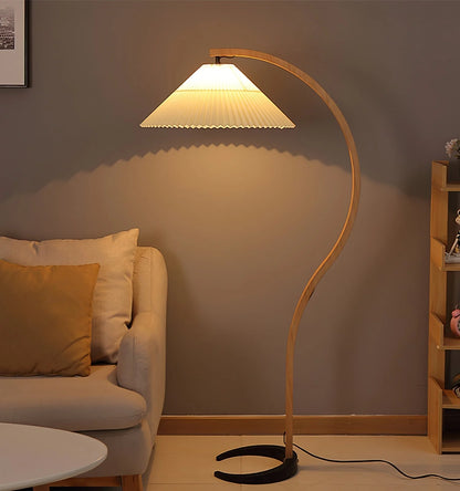 Nordic style floor lamp with a curved beech wood stand and a white pleated shade, casting a warm glow in a modern living room beside a neutral sofa.