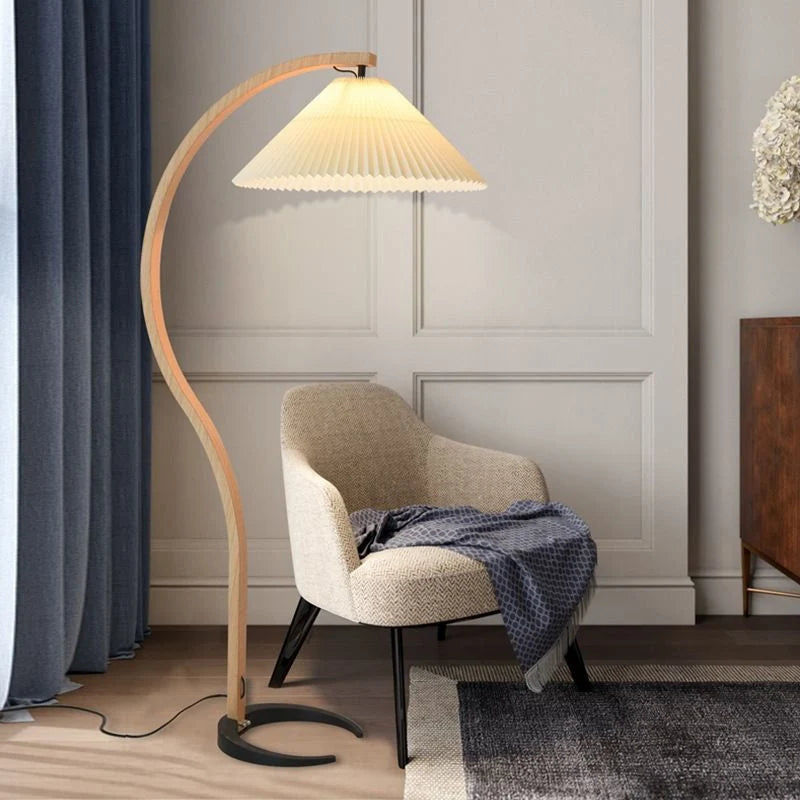 Nordic style floor lamp with a curved beech wood stand and a pleated fabric shade, positioned next to a modern armchair in a contemporary living room.