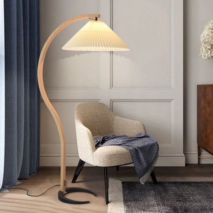 Nordic style floor lamp with a curved beech wood stand and a pleated fabric shade, positioned next to a modern armchair in a contemporary living room.