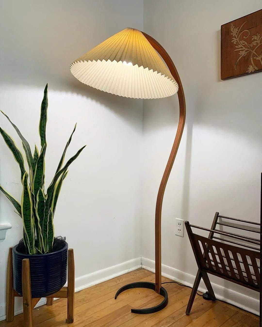 Nordic style floor lamp with a curved beech wood stand and a pleated shade, illuminating a corner next to a snake plant in a blue pot.