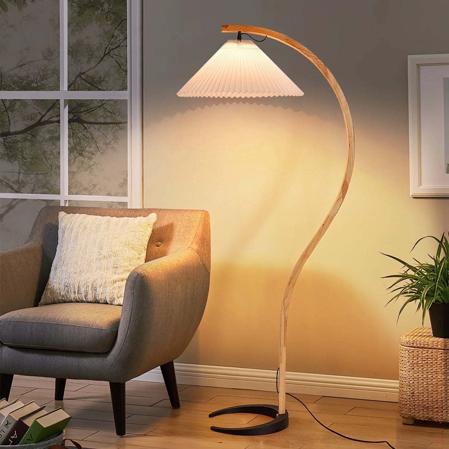 Nordic beech wood floor lamp with a curved stem and white pleated shade, casting a warm glow next to a grey armchair in a cosy living room.