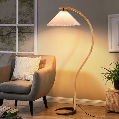 Nordic beech wood floor lamp with a curved stem and white pleated shade, casting a warm glow next to a grey armchair in a cosy living room.