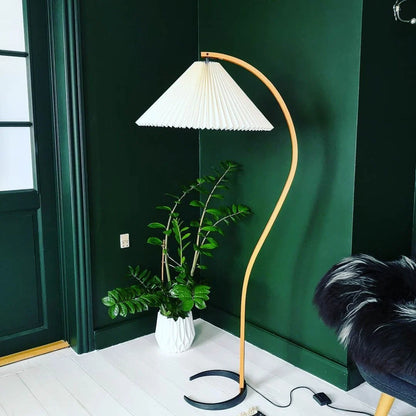 Nordic style floor lamp with a curved beech wood stand and a white pleated shade, positioned in a corner against a dark green wall next to a plant.