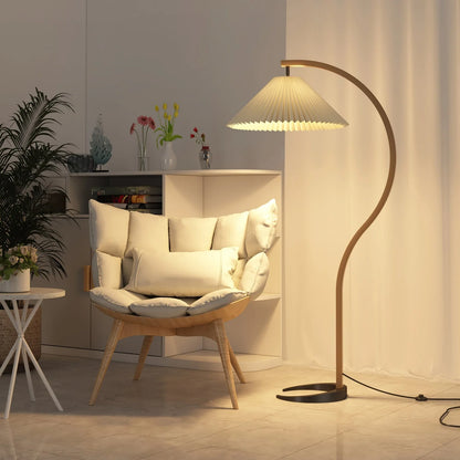 Nordic style floor lamp with a curved beech wood stand and beige pleated shade, casting a warm glow in a modern living room beside a cushioned chair.