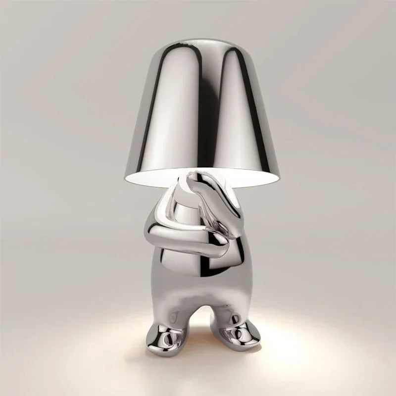 Modern silver rechargeable LED table lamp shaped like a small figure in a thoughtful pose with a touch-controlled light under a metallic shade.