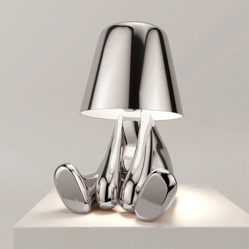 A modern silver rechargeable LED figure table lamp with a minimalist design, featuring a seated character shape and touch control on a white surface.