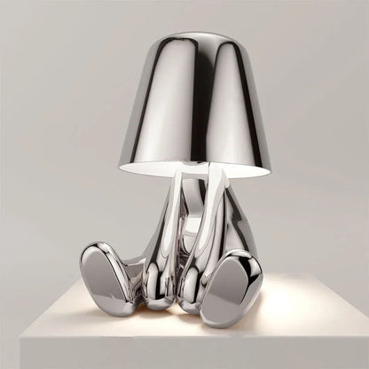 A modern silver rechargeable LED figure table lamp with a minimalist design, featuring a seated character shape and touch control on a white surface.