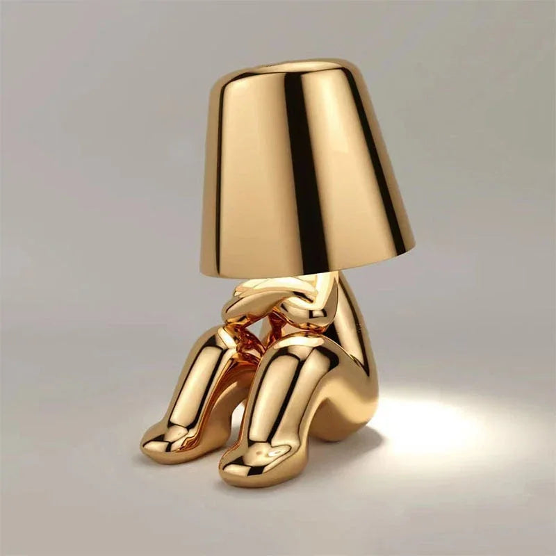 A modern gold rechargeable LED table lamp shaped like a seated figure with a minimalist lampshade, perfect for a bedside or desk setting.
