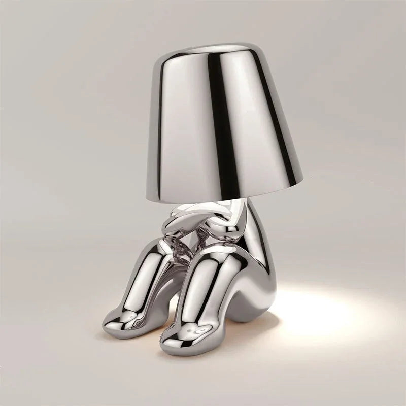 Modern rechargeable LED table lamp in a silver finish, featuring a minimalist figure design sitting with a lampshade head, perfect for bedside use.