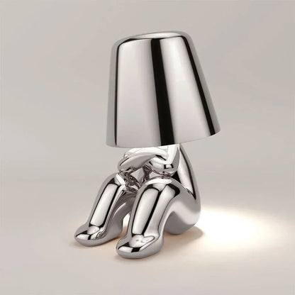 Modern rechargeable LED table lamp in a silver finish, featuring a minimalist figure design sitting with a lampshade head, perfect for bedside use.